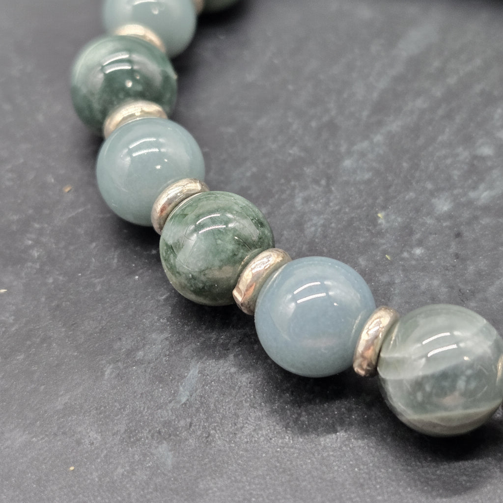 A close-up of a beaded strand featuring polished light blue-gray amazonite beads, dark green aventurine beads, and small metallic spacer beads, displayed on a dark slate surface. A watermark logo for 'luki lapidary' is visible in the bottom-left corner.