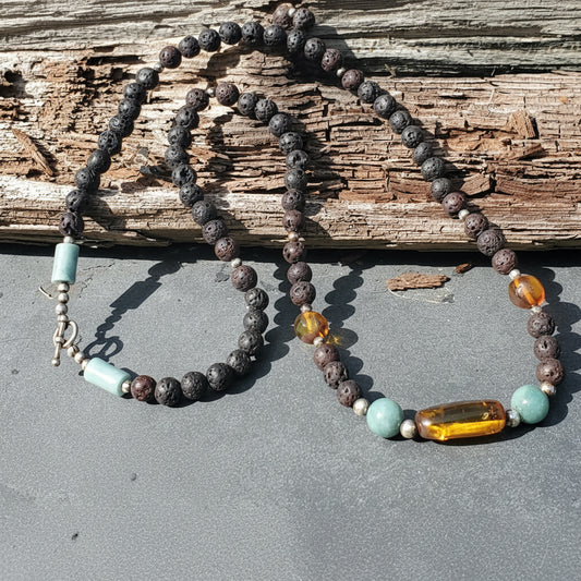 A beaded necklace with dark volcanic lava stone beads, light blue turquoise beads, and a large amber glass cylinder, displayed on a gray slate surface with a weathered wood background.