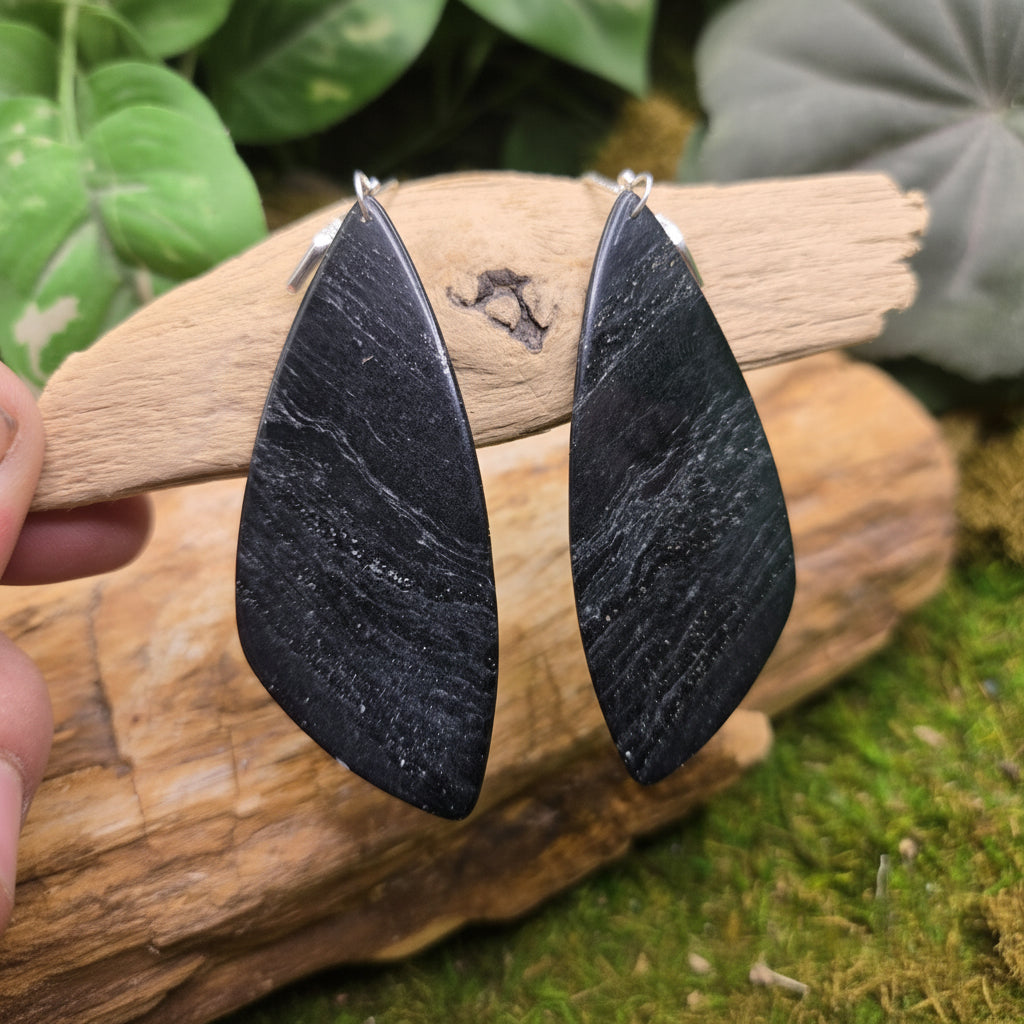 A pair of large, polished black stone teardrop earrings held by a hand against a natural background of wood and green moss. A watermark for 'luki lapidary' is visible in the bottom-left corner.