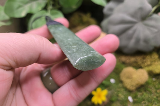 A hand holding a polished green jade pendant against a blurred natural background with leaves and moss. A watermark for 'luki lapidary' is visible in the lower-left corner.