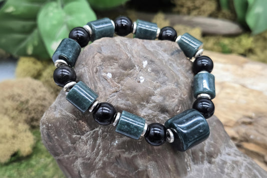 A beaded stretch bracelet displayed on a natural rock, featuring dark green rectangular labradorite beads and glossy black round beads separated by small silver spacer beads. The background shows blurred green foliage.