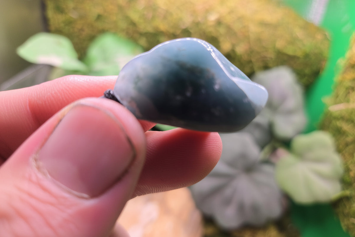 A hand holding a polished dark blue-gray stone with a glossy finish, set against a blurred background of green moss and foliage. A watermark for 'luki lapidary' is visible in the bottom-left corner.
