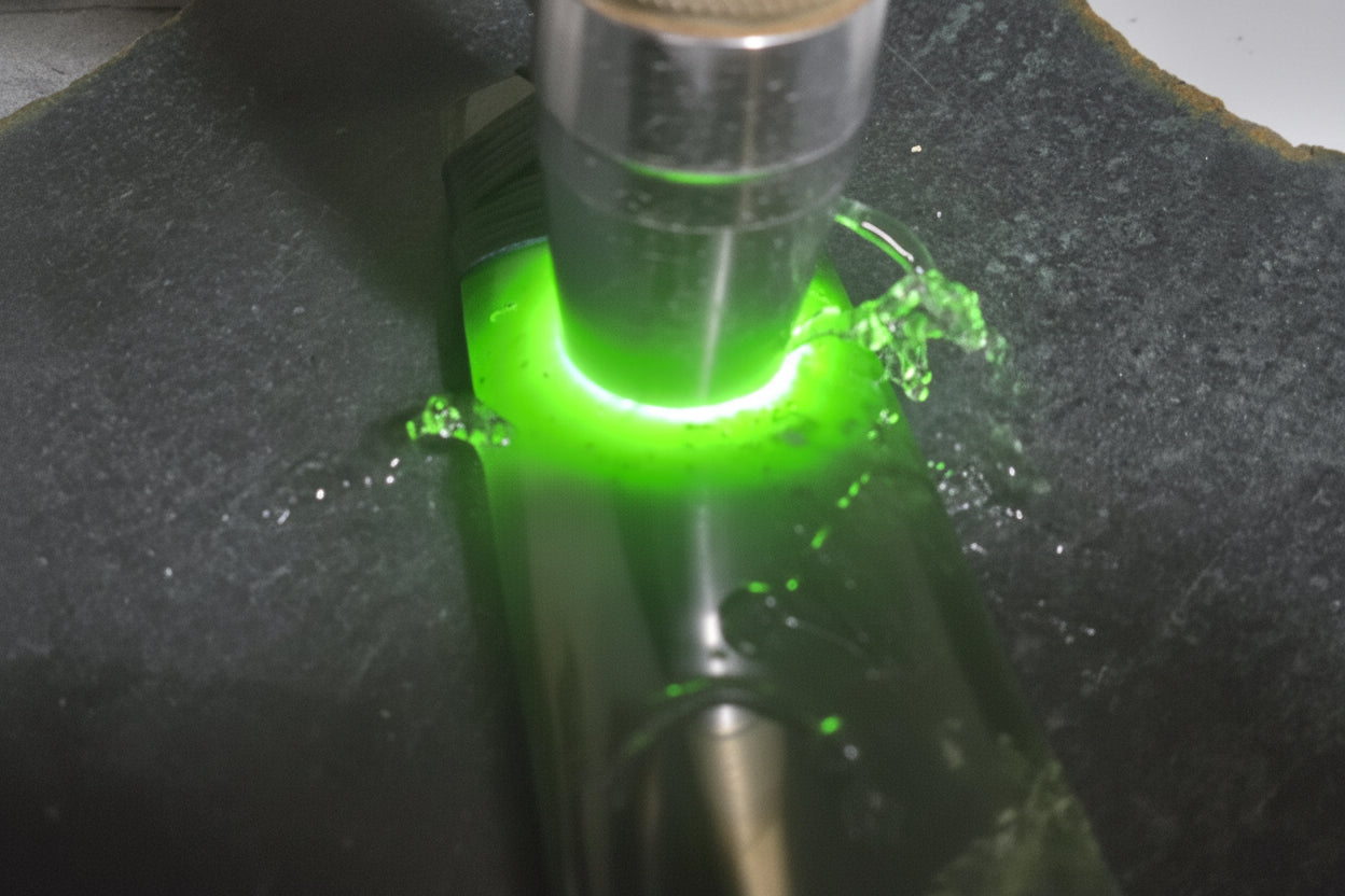 A close-up of a lapidary tool or stone sample under a metal cylinder, emitting a bright green light on a dark gray stone surface. A watermark reading 'luki lapidary artisan lapidary' is visible in the lower left.