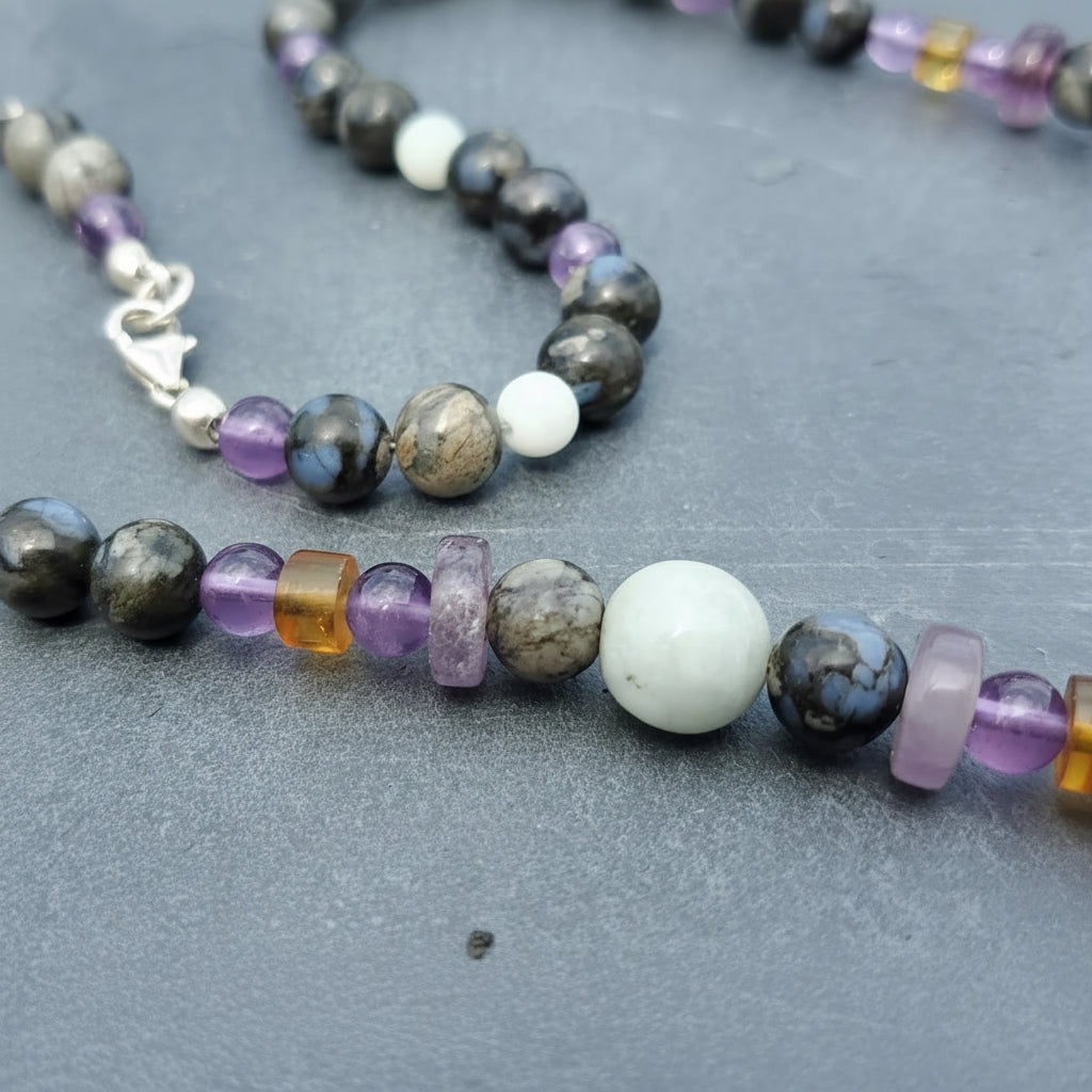 A close-up of a beaded jewelry strand with mixed gemstone beads in amethyst , black, jade, brown, and amber, laid on a gray textured surface. A silver clasp is visible on the left.
