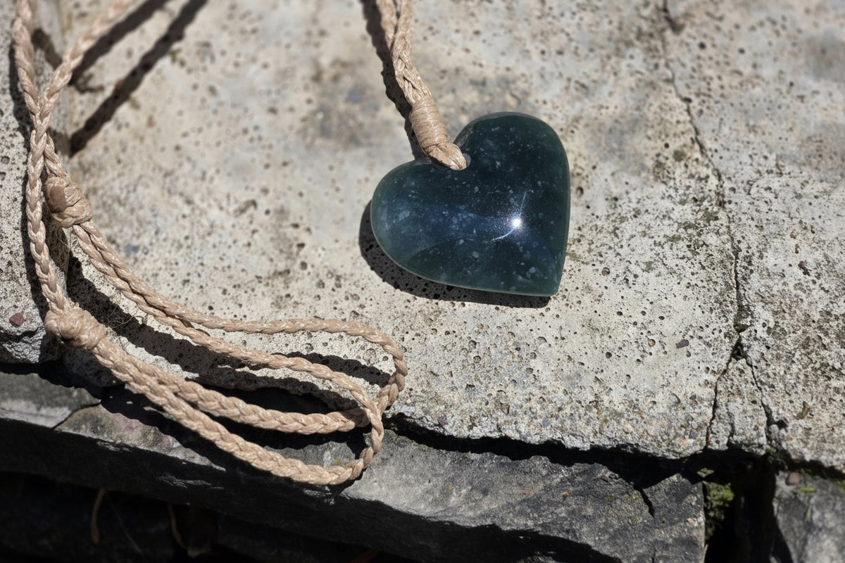 A heart-shaped polished dark blue stone pendant on a natural jute cord, resting on a textured gray stone surface.