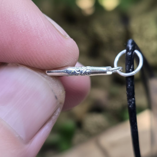 A close-up of a hand holding a small silver lapel pin with a ring attachment, shown against a blurred natural background. A 'luki lapidary' watermark is visible in the lower-left corner.