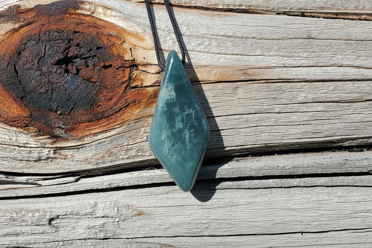 A polished teal-green teardrop-shaped gemstone pendant on a black cord, resting on weathered wooden planks next to a burnt wood section. A watermark for 'luki lapidary artisan lapidary' is visible in the bottom right corner.