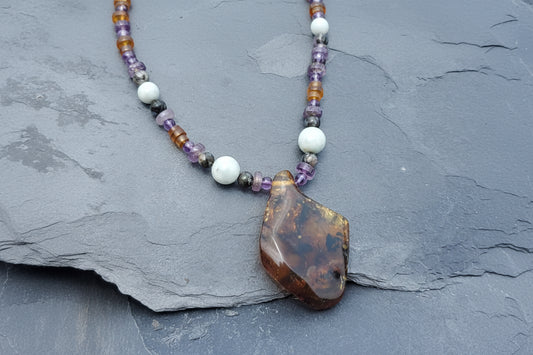 A handmade lapidary necklace featuring a polished brown agate pendant and white, purple, and amber beads, displayed on a dark gray slate surface.