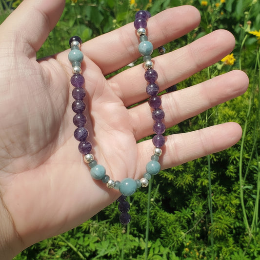 A beaded necklace with purple amethyst, light blue jade, and silver spacer beads held in a person's hand, with a blurred green garden background.