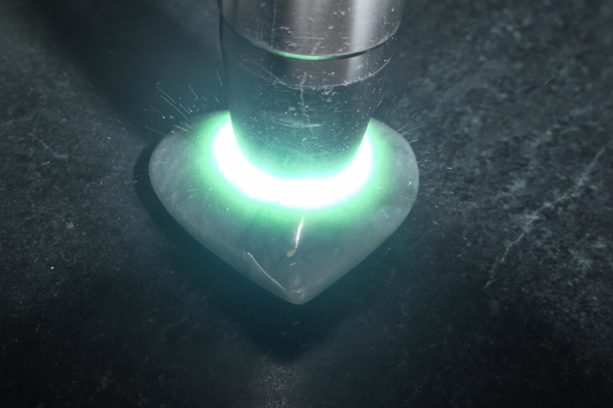 A close-up of a heart-shaped translucent stone or crystal placed under a metal lamp, glowing with a bright green light. The stone has a pointed tip and a smooth polished surface.