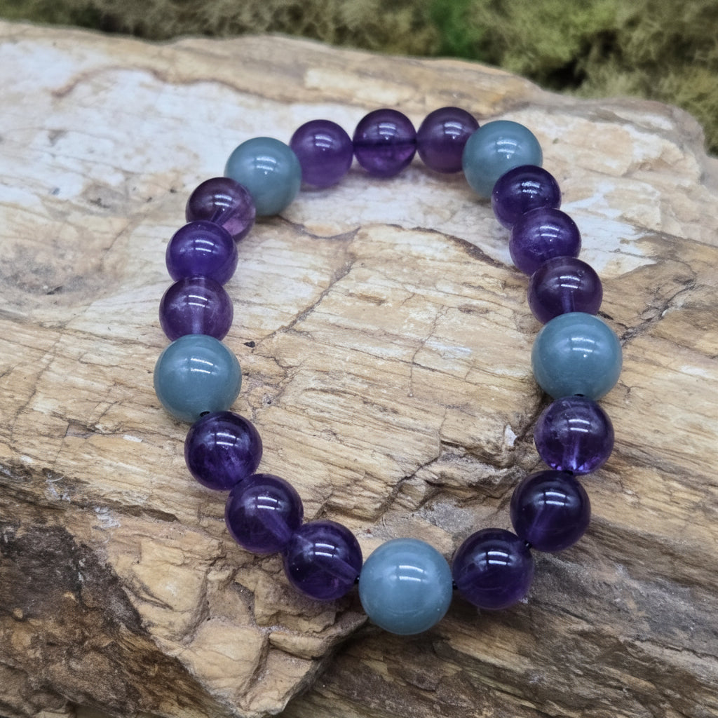 A beaded stretch bracelet made of alternating smooth purple amethyst and light blue labradorite beads, displayed on a textured natural rock with a blurred mossy background. A watermark logo for 'luki lapidary' is visible in the bottom-left corner.