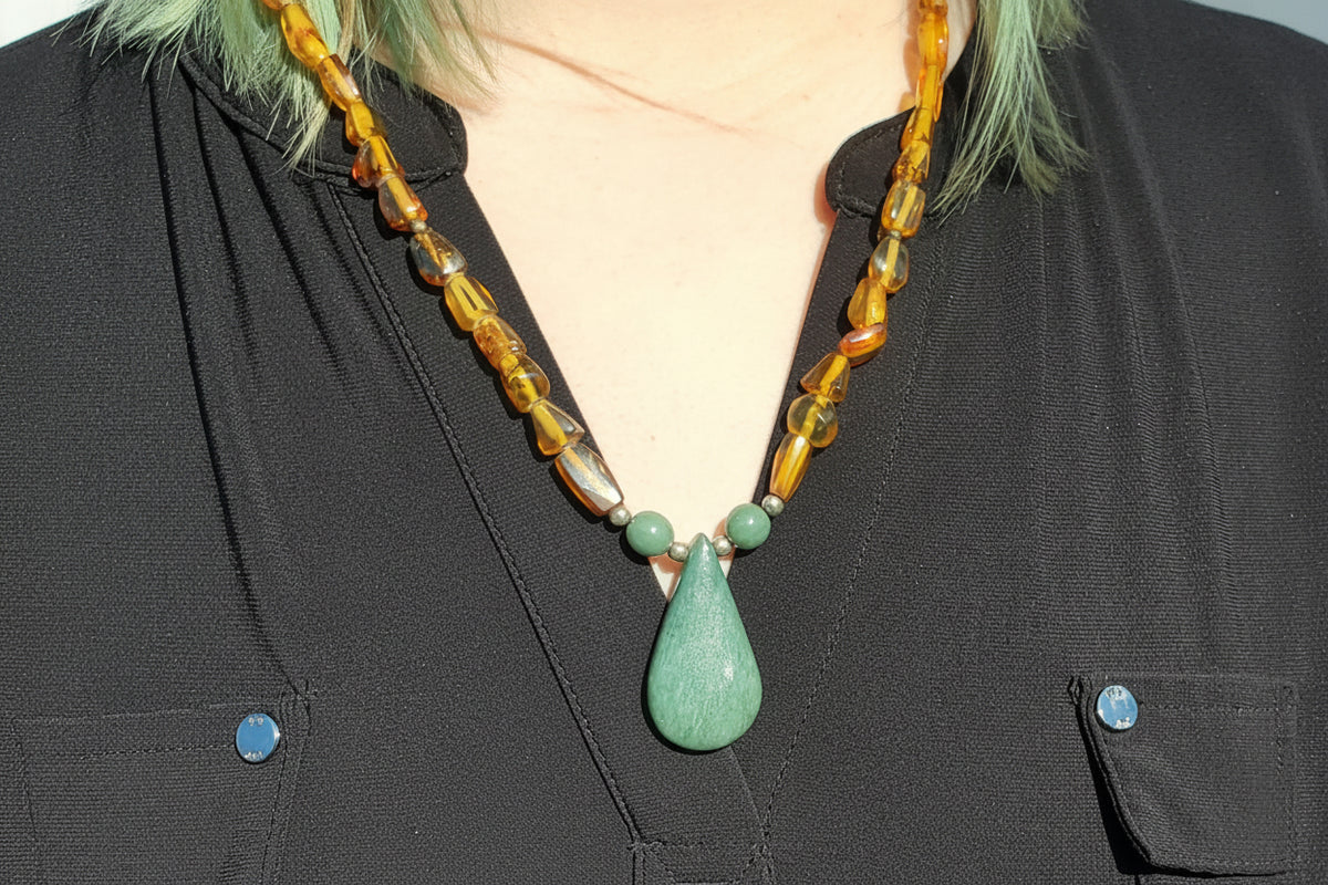A close-up of a person wearing a handmade necklace with a large green teardrop pendant, green round beads, and amber-colored translucent beads, styled with a black shirt. The logo 'luki lapidary' is visible in the bottom-left corner.