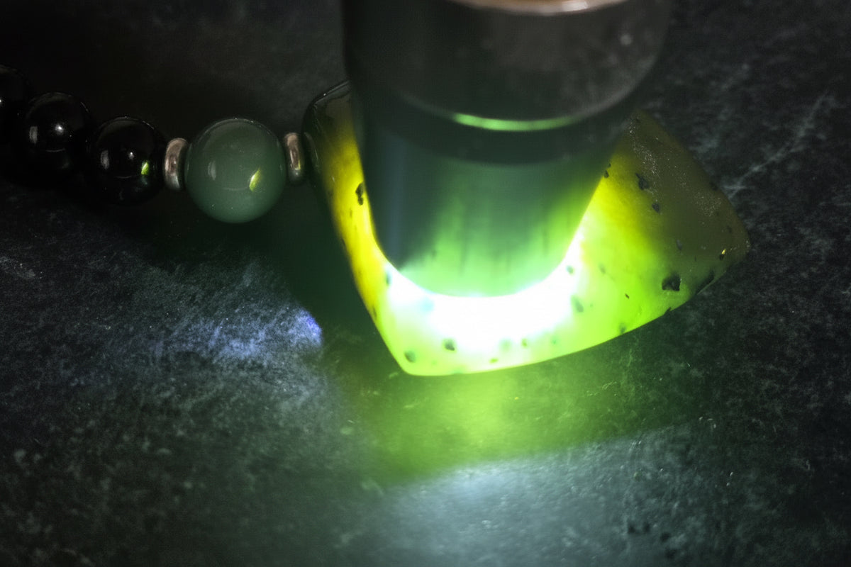 A close-up of a lapidary stone being illuminated by a flashlight, showing a bright green glow. Part of a beaded necklace with green beads is visible on the left.