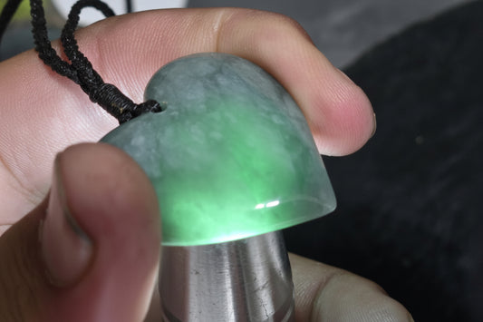 A close-up of a hand holding a heart-shaped green jade pendant with a black cord, positioned in front of a silver lapidary tool. A watermark for 'luki lapidary' is visible in the lower-left corner.
