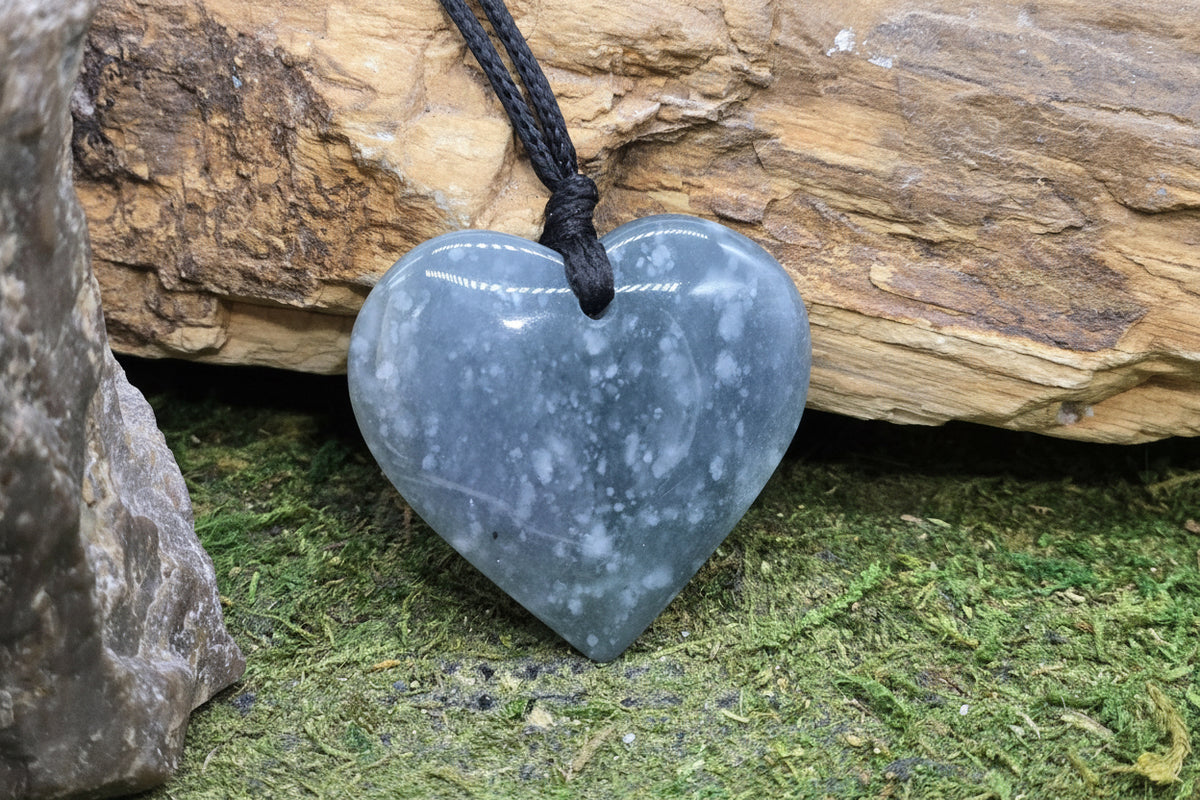 A heart-shaped polished gray stone pendant with white marbling, suspended from a black cord, resting against a natural rock and moss background. A 'luki lapidary' watermark is visible in the lower-left corner.