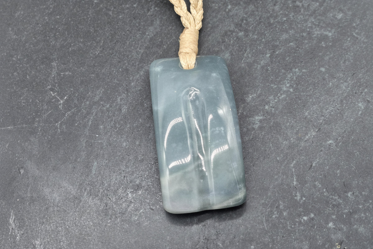 A rectangular carved stone pendant with a carved figure design, made of a translucent pale blue-gray material and suspended from a beige cord, shown on a dark gray textured background. A watermark logo for 'luki lapidary' is visible in the bottom-left corner.