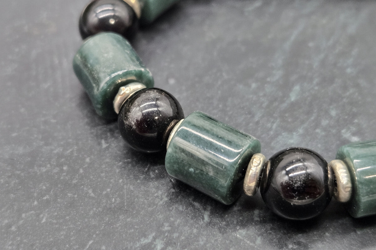 A close-up of a beaded strand featuring polished dark green cylindrical beads and glossy black spherical beads separated by small metallic spacer rings, set against a dark gray textured background.
