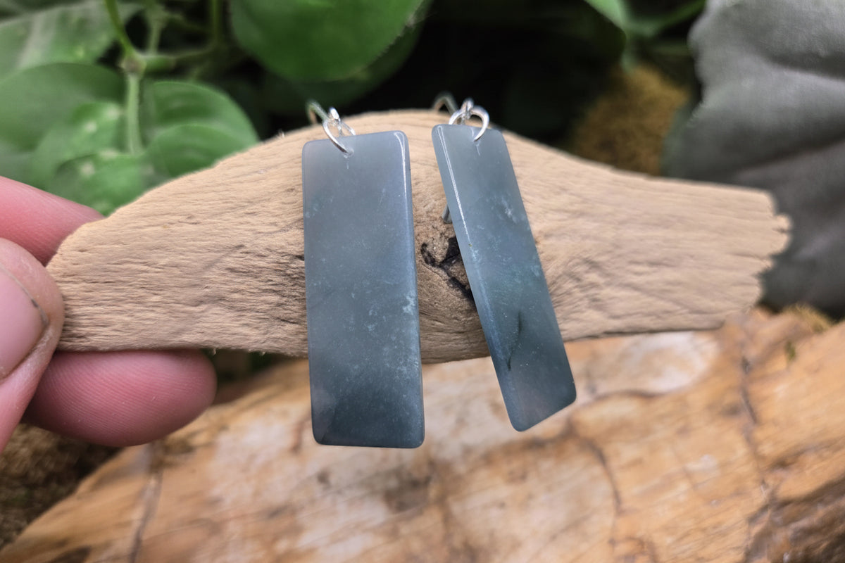 A pair of rectangular drop earrings made from dark gray polished stone, held on a piece of driftwood by a hand. The background features green foliage and a textured wooden surface, with a 'luki lapidary' watermark in the bottom-left corner.