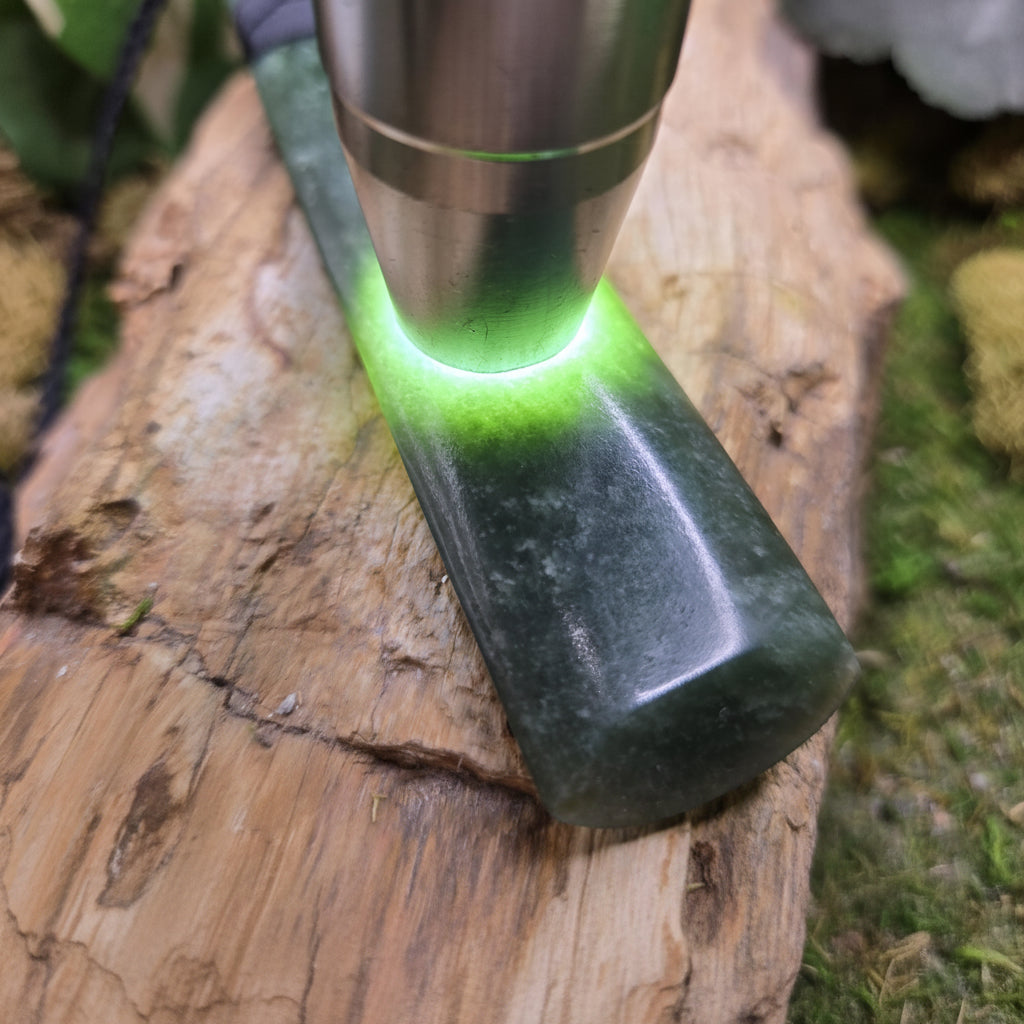 A close-up of a lapidary tool or light source shining green light onto a polished dark green stone block resting on a piece of wood, with a visible watermark reading 'luki lapidary' in the lower left.