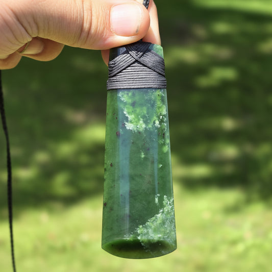 A hand holds a polished green stone pendant with a black cord wrap and black hanging cord, photographed outdoors against a blurred green background. A watermark reading 'luki lapidary artisan lapidary' appears in the lower left.
