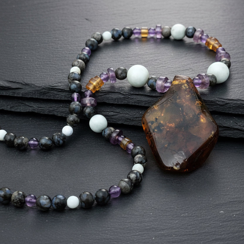 A beaded necklace with a large polished amber-brown stone pendant, displayed on a dark gray surface with a weathered wooden background. The beads include white, purple, gray, and amber-orange tones.