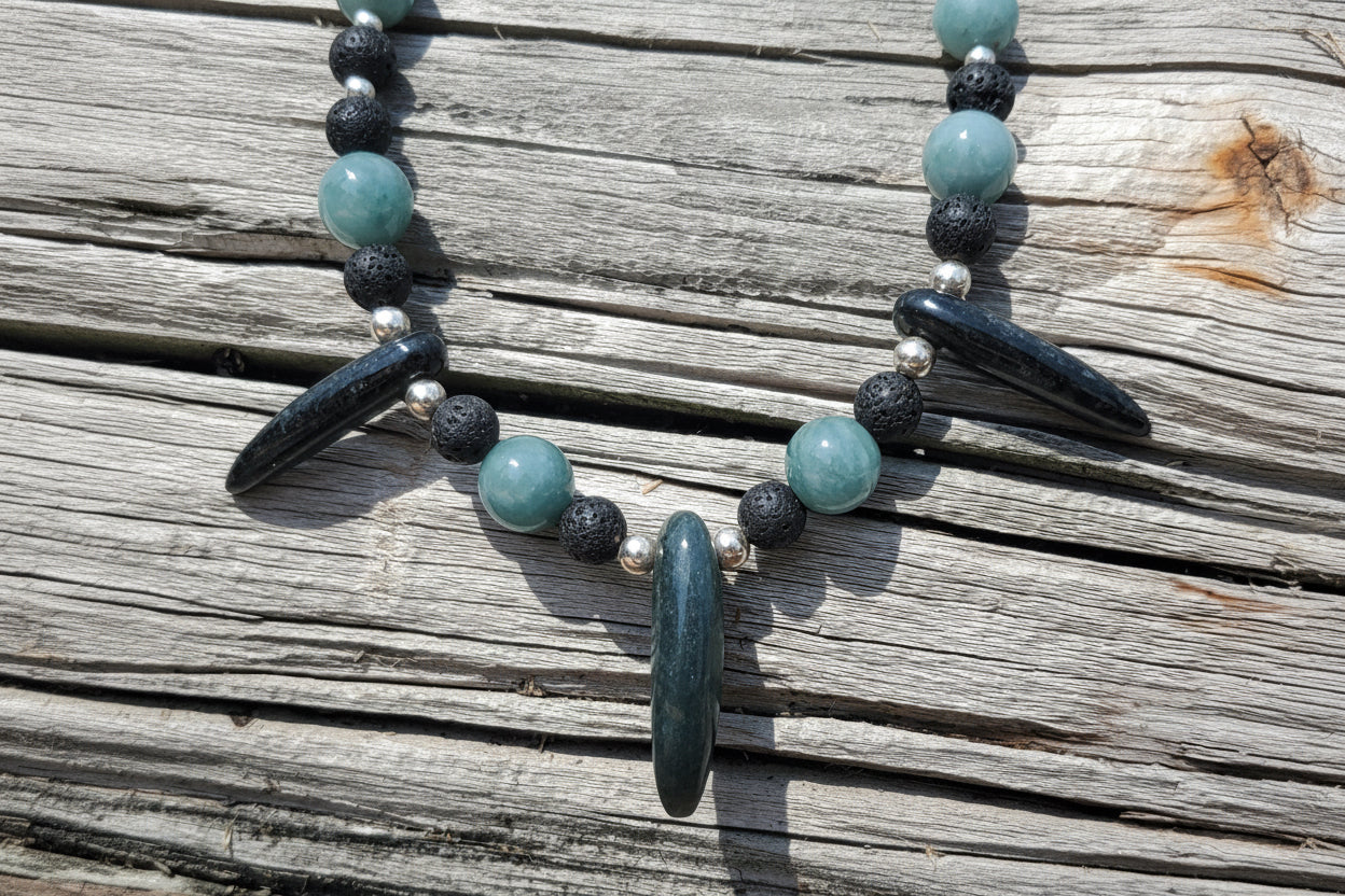 A beaded necklace with light blue-green round jade beads, black lava stone beads, silver spacer beads, and dark polished stone pendants, displayed on weathered gray wood planks. 