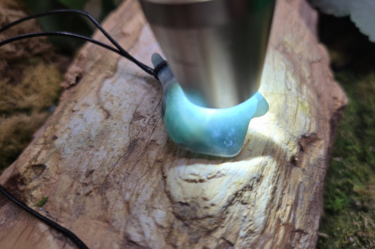 A close-up of a light blue-green translucent stone pendant or cabochon resting on a rough log, with a metal light source shining into it. The pendant has a black cord and is photographed outdoors on natural wood.
