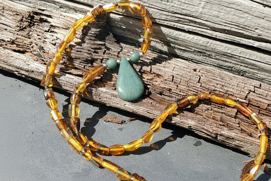 A beaded necklace with translucent amber-yellow and brown beads, featuring a central green teardrop pendant and two small green beads, displayed on weathered wood outdoors.