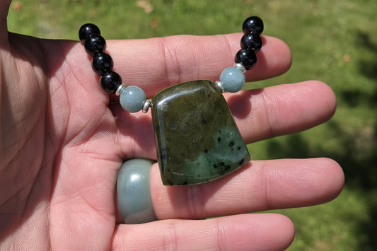 A hand holding a beaded necklace with a large polished green stone pendant, black round beads, and light blue accent beads, photographed outdoors with grass in the background.