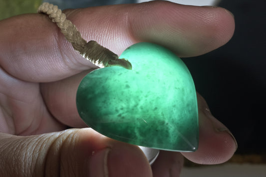 A heart-shaped green jade pendant with a natural fiber loop, held in a hand. The pendant has a mottled green and white marbled pattern.