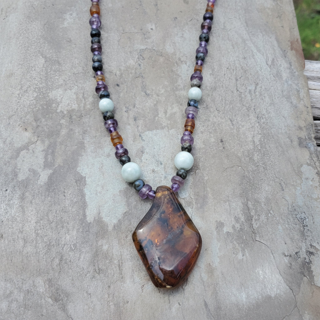 A beaded necklace with a large polished brown stone pendant, displayed on a textured gray stone surface. The necklace features a mix of purple amethyst beads, white Jade beads, and translucent amber beads.