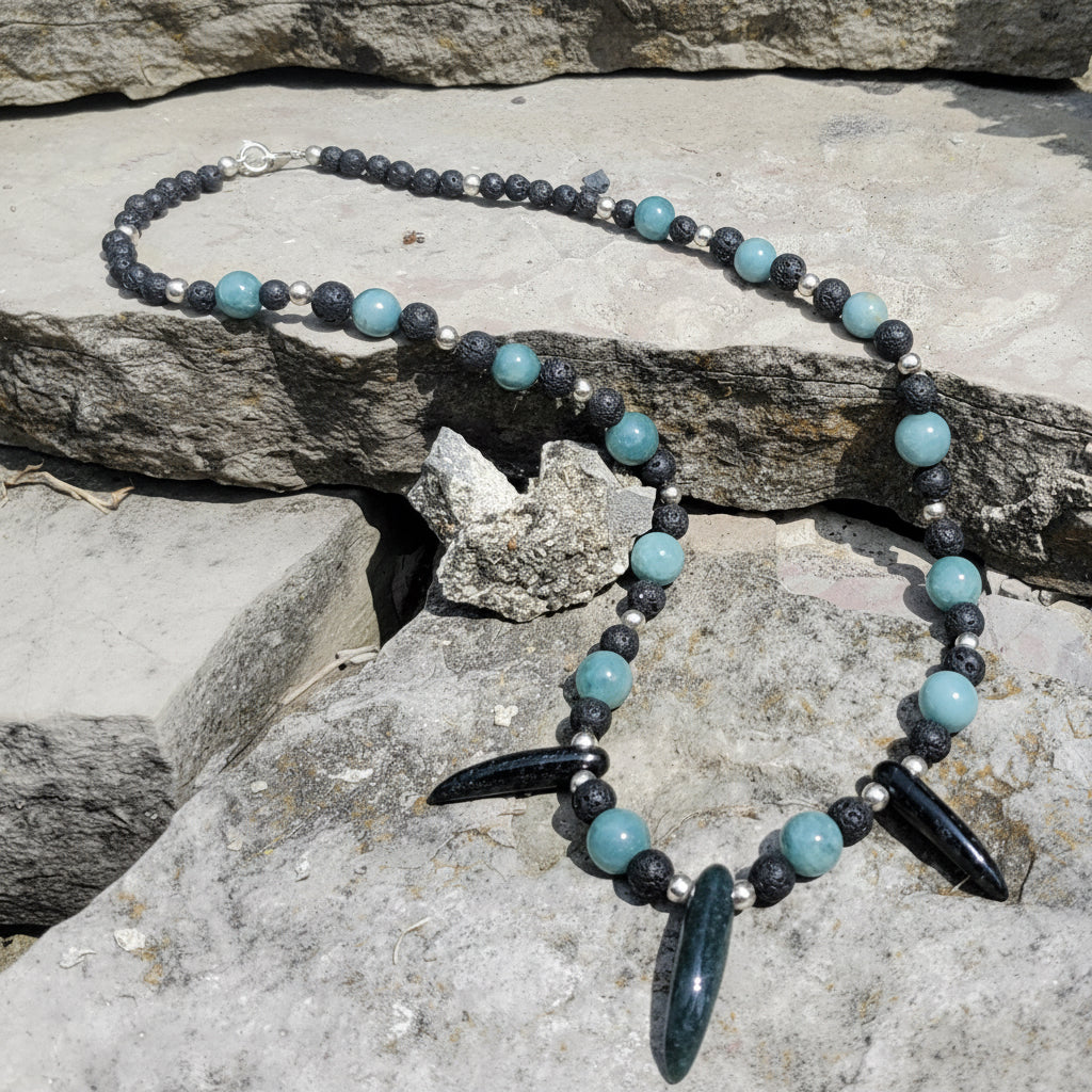 A beaded necklace with light blue and dark gray jade beads, featuring two large dark blue-green teardrop pendants, displayed on a gray stone surface. 