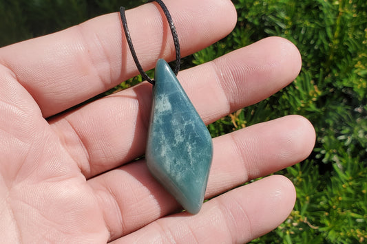 A hand holding a polished blue-green stone pendant on a black cord, with green foliage blurred in the background. A watermark reading 'luki lapidary artisan lapidary' is visible in the lower-left corner.