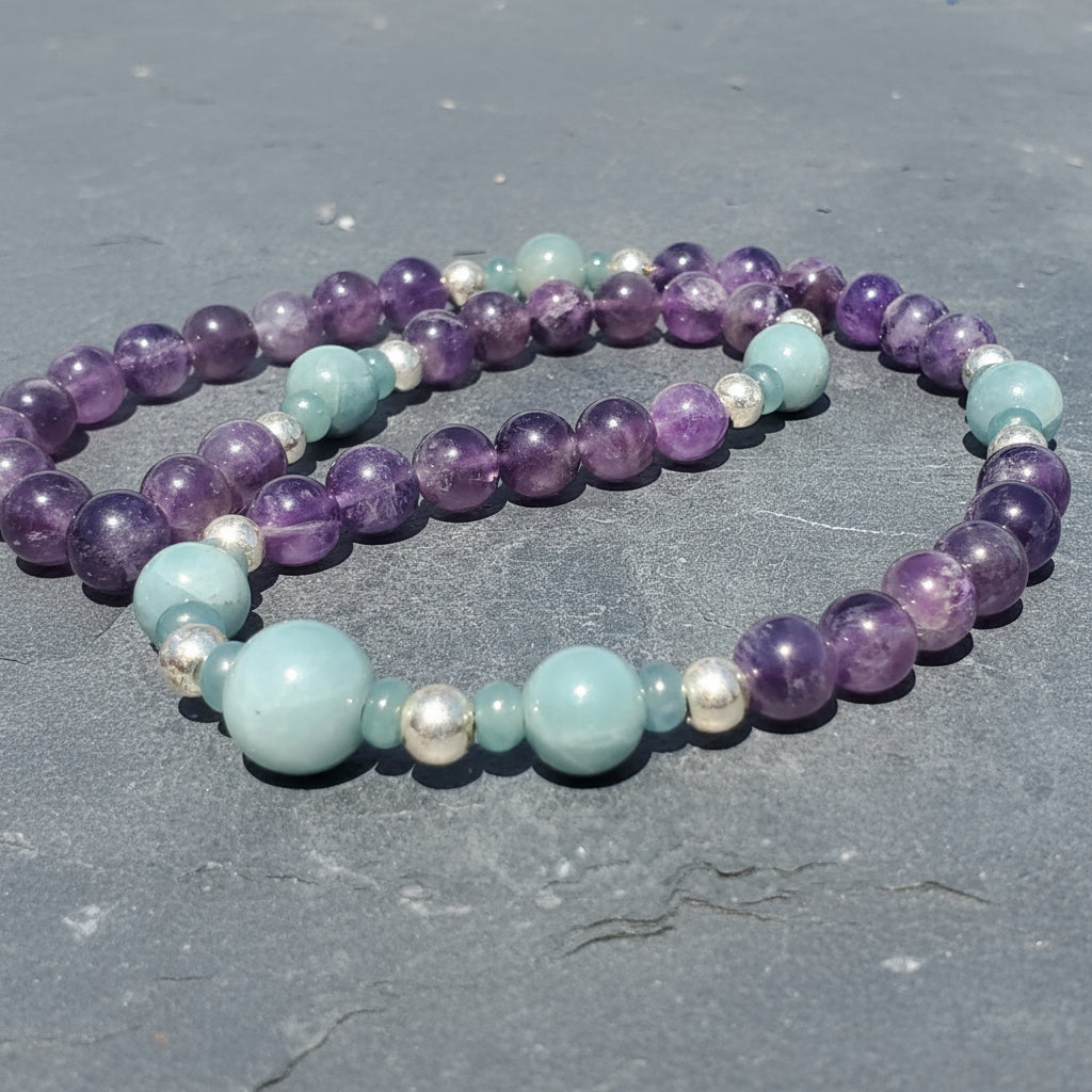 Two beaded stretch bracelets with purple amethyst, light blue jade, and small silver spacer beads, displayed on a gray stone surface. 