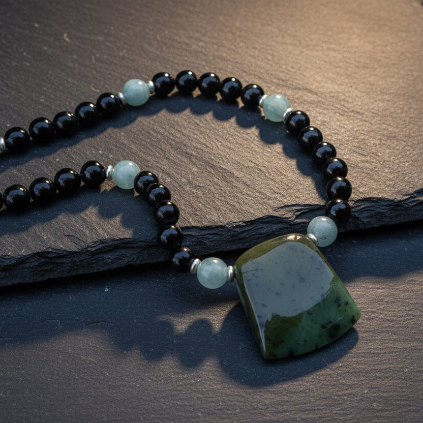 A beaded necklace with black round beads, light blue spacer beads, and a large polished green stone pendant, displayed on a textured gray stone surface with leaf shadows.