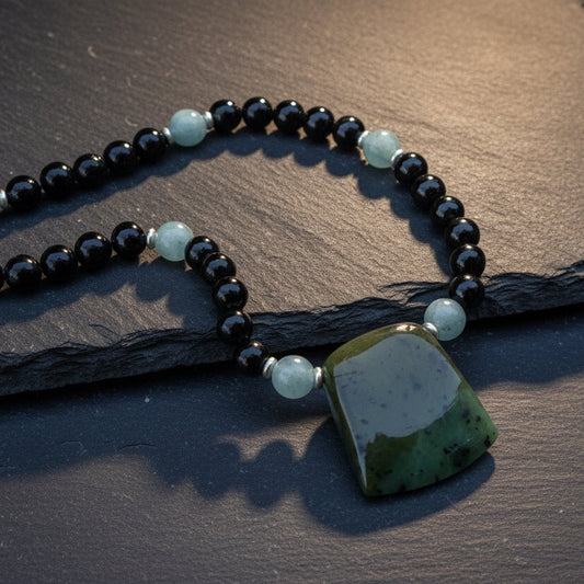 A beaded necklace with black round beads, light blue spacer beads, and a large polished green stone pendant, displayed on a textured gray stone surface with leaf shadows.