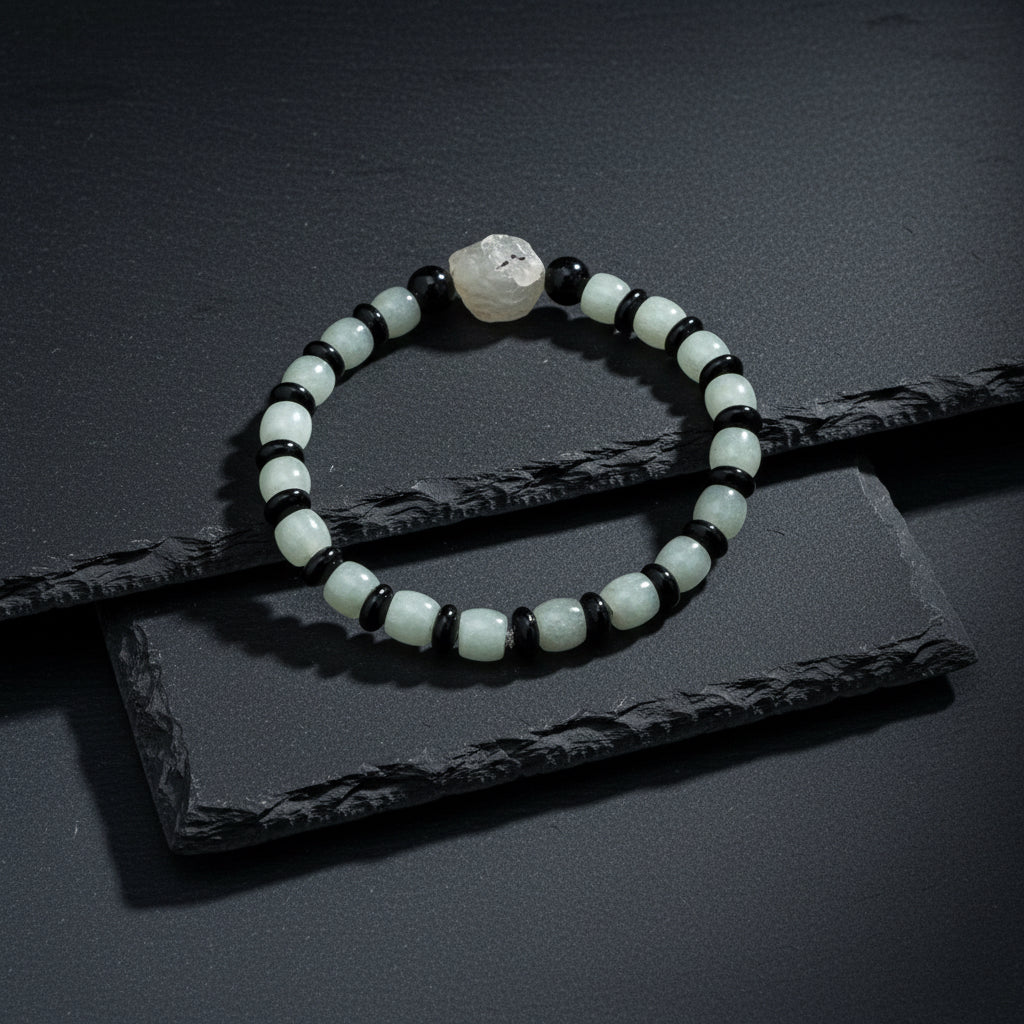 A beaded stretch bracelet featuring light green jade-like round beads, polished black oval beads, and a natural gray stone centerpiece, displayed on a textured gray wood background.