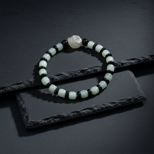 A beaded stretch bracelet featuring light green jade-like round beads, polished black oval beads, and a natural gray stone centerpiece, displayed on a textured gray wood background.
