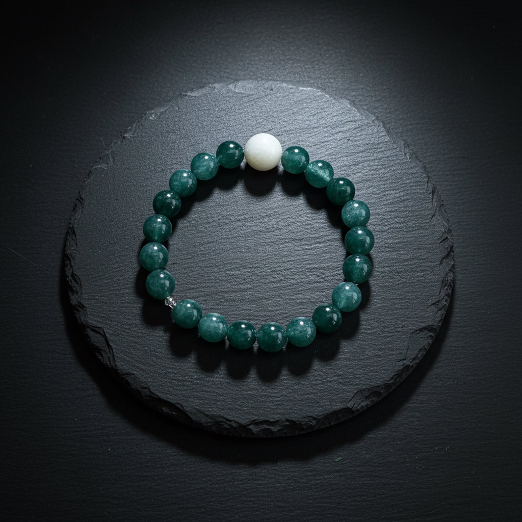 A beaded stretch bracelet made of faceted dark green gemstone beads and one smooth white bead, displayed on a weathered gray wood surface.