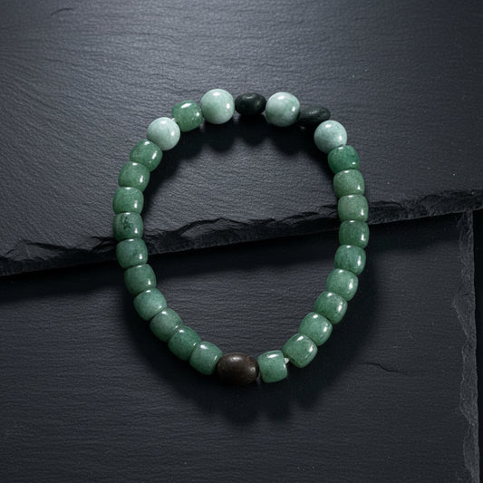A beaded stretch bracelet made of green jade beads with a few light blue, dark green, and black beads, displayed on a weathered gray wood surface.