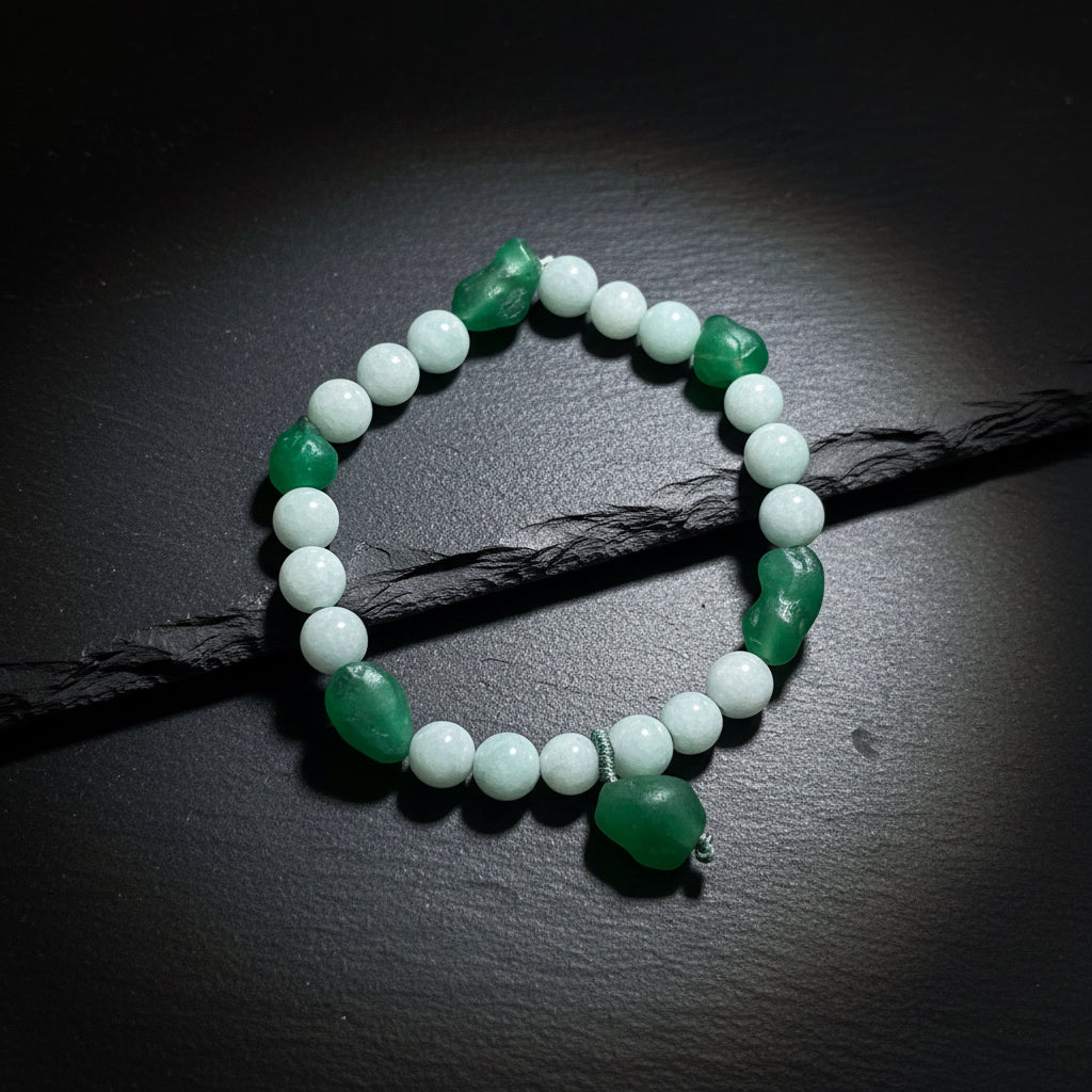 A beaded stretch bracelet featuring light gray-green round beads and dark green polished stone accents, displayed on a textured wooden surface.