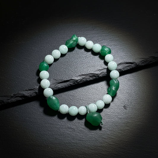 A beaded stretch bracelet featuring light gray-green round beads and dark green polished stone accents, displayed on a textured wooden surface.