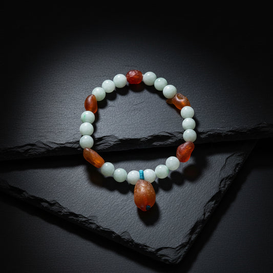 A beaded bracelet with light green jade-like round beads, reddish-brown polished accent beads, and a large teardrop-shaped brown bead dangling from a small blue cord, displayed on a weathered wood surface.