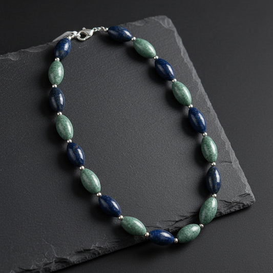 A beaded necklace with alternating dark navy blue and green stone beads, separated by small gold spacer rings, displayed on a rustic wooden surface.