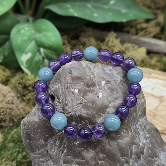 A beaded stretch bracelet made of alternating translucent purple amethyst and light blue turquoise beads, displayed on a gray stone with a green leaf in the blurred background. A watermark for 'luki lapidary artisan lapidary' is visible in the lower-left corner.