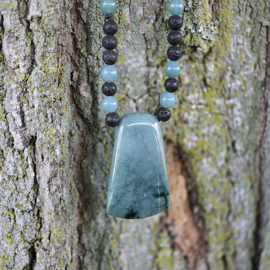 A polished light blue-green stone pendant necklace hanging against a textured tree trunk background, with a string of alternating pale blue and dark round beads above it. A watermark reading 'luki lapidary artisan lapidary' appears in the lower left.