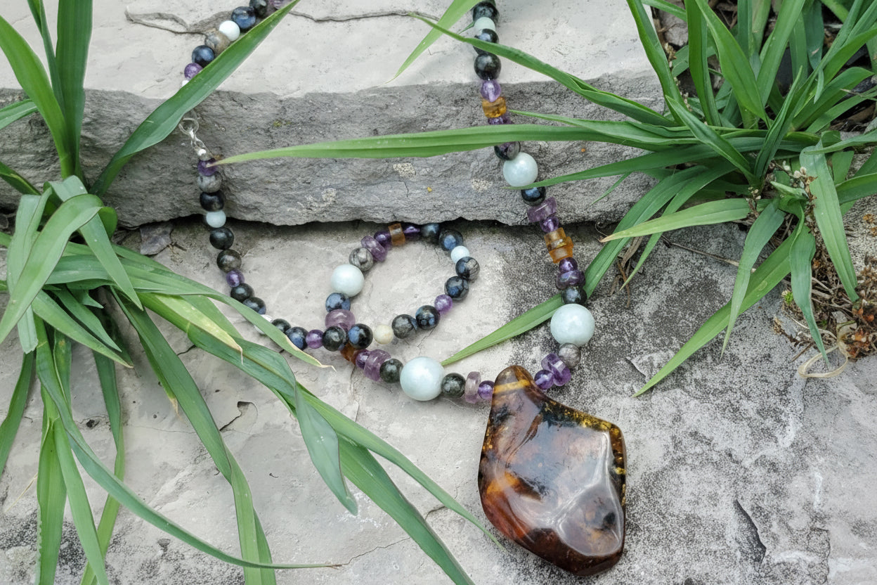A beaded necklace with a large polished amber  pendant and matching bead strands, arranged on gray stones with green grass. The necklace includes dark jade, amethyst, and amber beads.