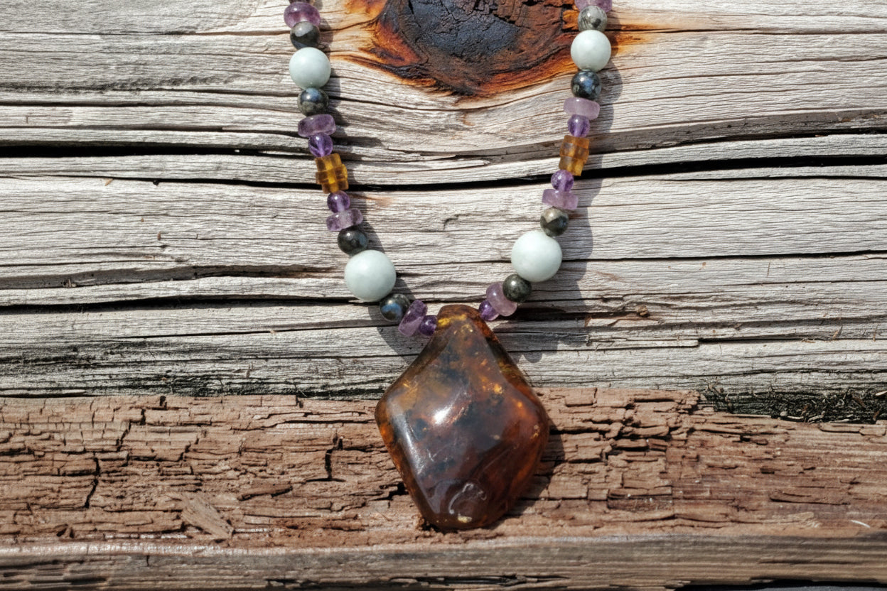 A handmade beaded necklace with a polished amber-brown irregular pendant, displayed on a weathered wooden surface. The necklace features a mix of jade, amethyst, and translucent amber beads.