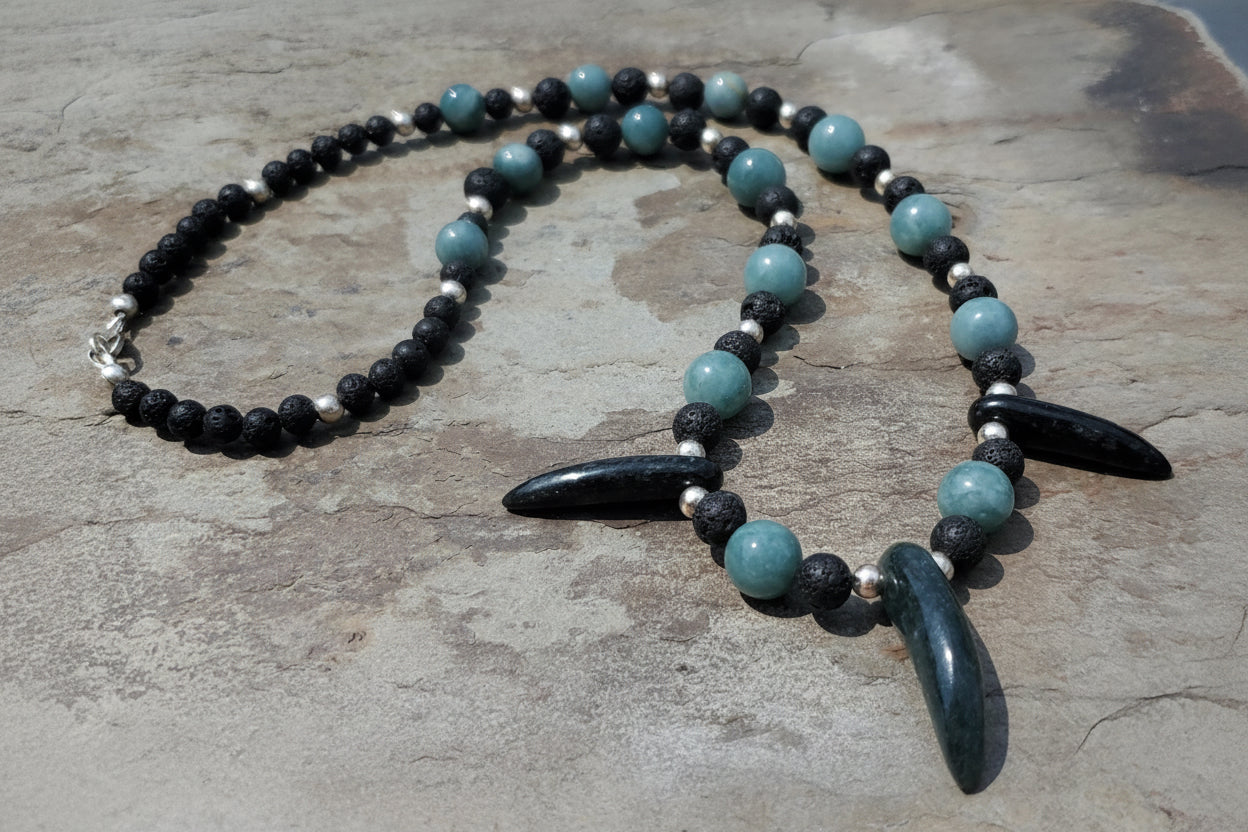 A beaded necklace with blue and black jade beads, two black horn-like pendants, and small silver spacer beads, displayed on a textured stone surface. 
