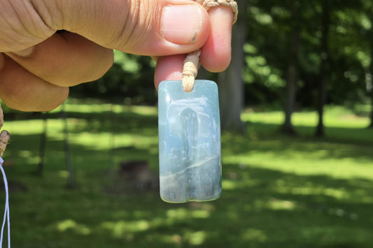 A hand holding a light blue carved stone pendant with a natural fiber cord, set against a blurred green outdoor background. A watermark for 'luki lapidary' is visible in the lower-left corner.